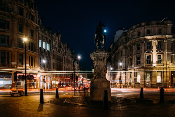An urban night scene featuring a busy street lined with historic buildings. Light trails from passing vehicles create vibrant streaks across the image, emanating a sense of movement. A prominent statue stands at the center, silhouetted against the dark sky, while streetlights cast a warm glow on the architecture.