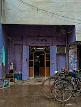 A quaint hair cutting salon with a purple exterior located in an alley. Two bicycles are parked in front of the salon, and a few household items like a stool and some containers are scattered around. The sign above the entrance reads 'Calcutta Hair Cutting Saloon,' and there are visible signs of wear and tear on the building.