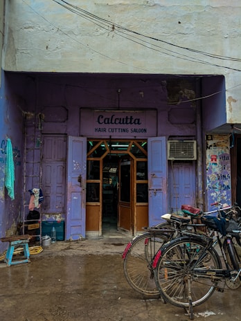 A quaint hair cutting salon with a purple exterior located in an alley. Two bicycles are parked in front of the salon, and a few household items like a stool and some containers are scattered around. The sign above the entrance reads 'Calcutta Hair Cutting Saloon,' and there are visible signs of wear and tear on the building.