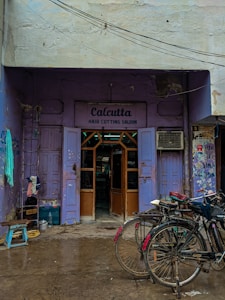 A quaint hair cutting salon with a purple exterior located in an alley. Two bicycles are parked in front of the salon, and a few household items like a stool and some containers are scattered around. The sign above the entrance reads 'Calcutta Hair Cutting Saloon,' and there are visible signs of wear and tear on the building.