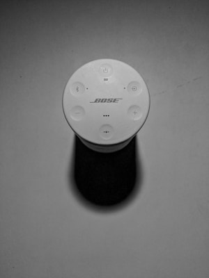 A top-down view of a Bose portable speaker, featuring its control buttons including power, volume, and Bluetooth icons. The speaker is casting a shadow, and the environment appears minimalistic with a smooth surface.