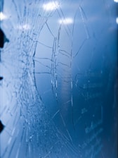 A close-up view of a cracked glass surface, displaying numerous fractures and intricate patterns against a blue background. The lighting highlights the texture and depth of the breaks.