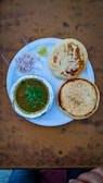 A plate containing two round, golden-brown breads and a serving of curry in a foil container. Garnished with green herbs, accompanied by chopped onions, two plastic spoons, and a wedge of lime. The setup is on a textured, brown table with hints of a pair of jeans and a hand at the bottom edge.
