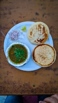 A plate containing two round, golden-brown breads and a serving of curry in a foil container. Garnished with green herbs, accompanied by chopped onions, two plastic spoons, and a wedge of lime. The setup is on a textured, brown table with hints of a pair of jeans and a hand at the bottom edge.