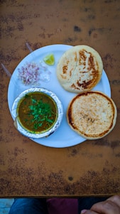 A plate containing two round, golden-brown breads and a serving of curry in a foil container. Garnished with green herbs, accompanied by chopped onions, two plastic spoons, and a wedge of lime. The setup is on a textured, brown table with hints of a pair of jeans and a hand at the bottom edge.