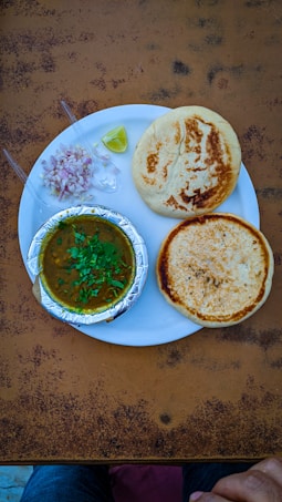 A plate containing two round, golden-brown breads and a serving of curry in a foil container. Garnished with green herbs, accompanied by chopped onions, two plastic spoons, and a wedge of lime. The setup is on a textured, brown table with hints of a pair of jeans and a hand at the bottom edge.
