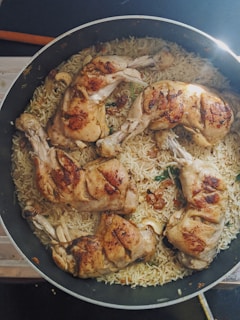 A large pan contains cooked chicken legs resting on a bed of seasoned rice. The chicken is golden brown, and the rice is intermixed with herbs and spices. The dish appears to be freshly cooked and is placed on a countertop.