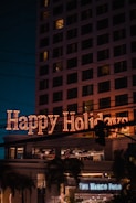 A festive neon sign displaying 'Happy Holidays' is prominently illuminated on a building, with multiple windows visible. Below, a sign for 'The Marco Polo' is noticeable, suggesting a hotel or business establishment. Palm trees and streetlights enhance the scene, while the sky is dark, indicating nighttime.