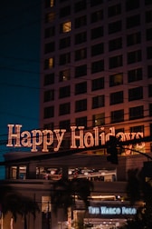 A festive neon sign displaying 'Happy Holidays' is prominently illuminated on a building, with multiple windows visible. Below, a sign for 'The Marco Polo' is noticeable, suggesting a hotel or business establishment. Palm trees and streetlights enhance the scene, while the sky is dark, indicating nighttime.