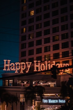 A festive neon sign displaying 'Happy Holidays' is prominently illuminated on a building, with multiple windows visible. Below, a sign for 'The Marco Polo' is noticeable, suggesting a hotel or business establishment. Palm trees and streetlights enhance the scene, while the sky is dark, indicating nighttime.