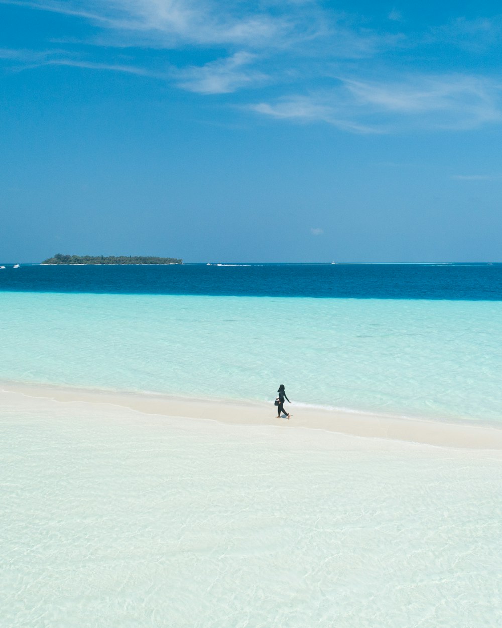person walking on beach viewing island under blue and white sky