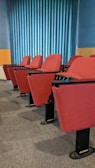 A row of red cushioned auditorium chairs with black metal bases is positioned on a gray carpet. Behind the chairs is a wall with vertical blue stripes, and the scene is well-lit with natural light.