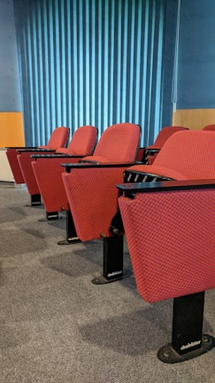 A row of red cushioned auditorium chairs with black metal bases is positioned on a gray carpet. Behind the chairs is a wall with vertical blue stripes, and the scene is well-lit with natural light.