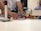 A person is assembling a construction made of colorful interlocking bricks on a table. Their hand is adjusting pieces, which include structures and various items like a small model house and poles. The background is slightly out of focus with blurred figures of people.