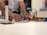 A person is assembling a construction made of colorful interlocking bricks on a table. Their hand is adjusting pieces, which include structures and various items like a small model house and poles. The background is slightly out of focus with blurred figures of people.