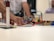 A person is assembling a construction made of colorful interlocking bricks on a table. Their hand is adjusting pieces, which include structures and various items like a small model house and poles. The background is slightly out of focus with blurred figures of people.