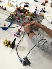 A detailed Lego setup is being assembled with various blocks and figurines. Two hands are actively engaging in arranging cables or tubes across the setup. The scene features structures like a ladder, several figurine characters, and a vehicle. Various colors and components stand out, adding a playful and creative element to the scene.