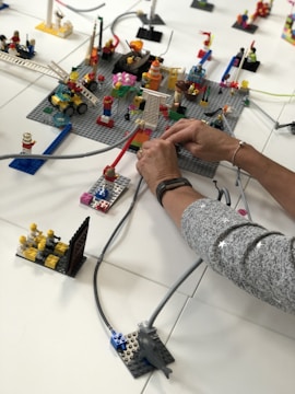 A detailed Lego setup is being assembled with various blocks and figurines. Two hands are actively engaging in arranging cables or tubes across the setup. The scene features structures like a ladder, several figurine characters, and a vehicle. Various colors and components stand out, adding a playful and creative element to the scene.