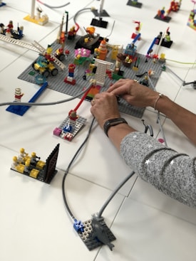 A detailed Lego setup is being assembled with various blocks and figurines. Two hands are actively engaging in arranging cables or tubes across the setup. The scene features structures like a ladder, several figurine characters, and a vehicle. Various colors and components stand out, adding a playful and creative element to the scene.