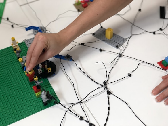 A hand is placing figures on a Lego platform surrounded by a network of black wires laid out on a white table. Several Lego-like figures and blocks are positioned on the green base, showcasing an assembly of small, colorful constructions.
