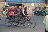 An older man rides a bicycle rickshaw on a busy street, with others in the background on bicycles and a motorbike. The rickshaw is red and blue, with an empty passenger seat, and there are auto-rickshaws parked nearby. The street is bordered by a stone wall and topped with blue metal fencing.