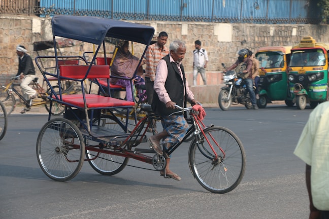 An older man rides a bicycle rickshaw on a busy street, with others in the background on bicycles and a motorbike. The rickshaw is red and blue, with an empty passenger seat, and there are auto-rickshaws parked nearby. The street is bordered by a stone wall and topped with blue metal fencing.