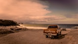 A vintage Toyota pickup truck is parked on a sandy beach beside the ocean. The vehicle faces away from the camera, with its logo clearly visible on the tailgate. The scene features rugged coastal rocks on the left side and the ocean stretching into the horizon on the right. The sky is filled with thick clouds casting a warm, sepia-toned light over the entire scene.