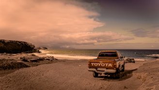 A vintage Toyota pickup truck is parked on a sandy beach beside the ocean. The vehicle faces away from the camera, with its logo clearly visible on the tailgate. The scene features rugged coastal rocks on the left side and the ocean stretching into the horizon on the right. The sky is filled with thick clouds casting a warm, sepia-toned light over the entire scene.