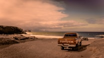 A vintage Toyota pickup truck is parked on a sandy beach beside the ocean. The vehicle faces away from the camera, with its logo clearly visible on the tailgate. The scene features rugged coastal rocks on the left side and the ocean stretching into the horizon on the right. The sky is filled with thick clouds casting a warm, sepia-toned light over the entire scene.