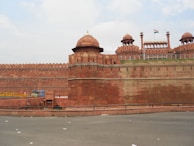 An expansive view of a historic fort with large red sandstone walls, topped by domed structures and embellished with intricate architectural details. The Indian national flag is visible atop one of the buildings. A few people are seen walking near the entrance, and the sky appears partially cloudy.