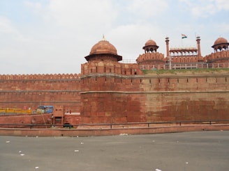 An expansive view of a historic fort with large red sandstone walls, topped by domed structures and embellished with intricate architectural details. The Indian national flag is visible atop one of the buildings. A few people are seen walking near the entrance, and the sky appears partially cloudy.