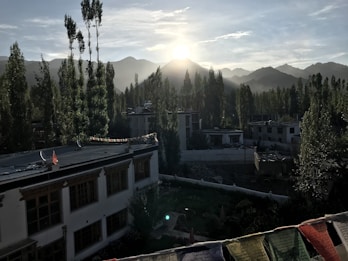 A picturesque landscape featuring residential buildings surrounded by tall trees, with mountains in the background. The sun is rising or setting over the mountains, casting a serene glow. Colorful prayer flags are visible in the foreground, adding a cultural element to the scene.