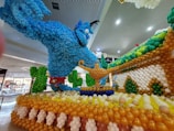 A large and colorful balloon sculpture depicting a blue genie emerging from a magic lamp, surrounded by decorative elements crafted from balloons. The display is located in a shopping area with visible signage.