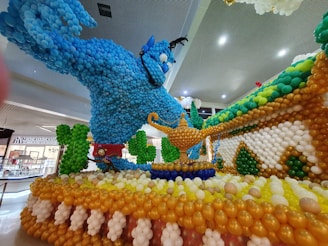 A large and colorful balloon sculpture depicting a blue genie emerging from a magic lamp, surrounded by decorative elements crafted from balloons. The display is located in a shopping area with visible signage.