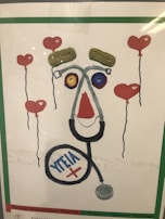 A creative artwork features elements resembling a face made from various objects, including a stethoscope forming the outline, yarn or string for the eyebrows, and colorful circular objects for the eyes. Surrounding the face are floating heart-shaped balloons with red color as the main theme.