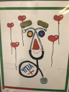 A creative artwork features elements resembling a face made from various objects, including a stethoscope forming the outline, yarn or string for the eyebrows, and colorful circular objects for the eyes. Surrounding the face are floating heart-shaped balloons with red color as the main theme.