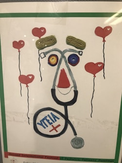 A creative artwork features elements resembling a face made from various objects, including a stethoscope forming the outline, yarn or string for the eyebrows, and colorful circular objects for the eyes. Surrounding the face are floating heart-shaped balloons with red color as the main theme.