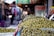 A market stall filled with a large quantity of green olives displayed in trays. The background features a bustling market scene with blurred figures of people walking and colorful items hanging or stacked. The atmosphere suggests a busy and lively marketplace.