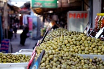 A market stall filled with a large quantity of green olives displayed in trays. The background features a bustling market scene with blurred figures of people walking and colorful items hanging or stacked. The atmosphere suggests a busy and lively marketplace.