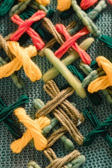 Various thick yarn threads are interwoven in an intricate pattern on a textured fabric. The threads form a repetitive cross-stitched design in multiple colors such as red, yellow, brown, green, and olive. The base fabric appears to have a woven structure, providing a contrast to the vibrant, soft yarns.