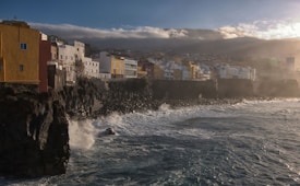 A coastline with a rough ocean and waves crashing against dark cliffs, lined with colorful buildings. The buildings are mostly white, yellow, and earthy tones, creating a striking contrast against the rocks. The sky is cloudy with a hint of sunlight breaking through, casting a soft glow over the scene.