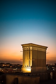 An ancient wind tower stands prominently against a vibrant sunset sky, casting a warm glow over the surrounding traditional buildings. The structure features tall, vertical slits for ventilation, characteristic of Persian architecture, and is highlighted by artificial lights at its base.