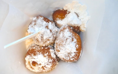 Four small doughnuts are covered with a generous dusting of powdered sugar. They are placed inside a white paper bag, with a skewer sticking out from the side.