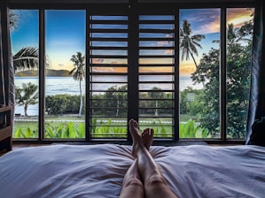 A view from a bed looking out through large windows at a tropical beach scene. The foreground shows a pair of relaxed legs resting on a white bed against the backdrop of lush greenery, palm trees, and the ocean. The sky is beautifully lit with colors of sunset or sunrise.