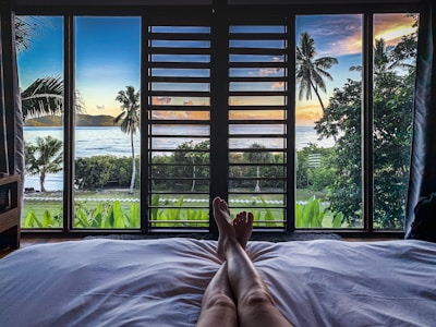 A view from a bed looking out through large windows at a tropical beach scene. The foreground shows a pair of relaxed legs resting on a white bed against the backdrop of lush greenery, palm trees, and the ocean. The sky is beautifully lit with colors of sunset or sunrise.