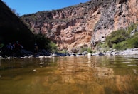 A clear mountain stream flows gently through a canyon with rocky, rugged cliffs. On the left side, a group of people wearing outdoor gear stand by the water's edge, some with bicycles. The sunlight casts bright and dark shadows across the landscape, highlighting the earthy tones of the rocks and the greens of the surrounding vegetation.