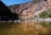 A clear mountain stream flows gently through a canyon with rocky, rugged cliffs. On the left side, a group of people wearing outdoor gear stand by the water's edge, some with bicycles. The sunlight casts bright and dark shadows across the landscape, highlighting the earthy tones of the rocks and the greens of the surrounding vegetation.