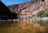 A clear mountain stream flows gently through a canyon with rocky, rugged cliffs. On the left side, a group of people wearing outdoor gear stand by the water's edge, some with bicycles. The sunlight casts bright and dark shadows across the landscape, highlighting the earthy tones of the rocks and the greens of the surrounding vegetation.