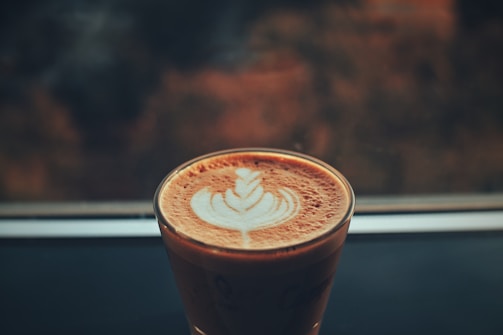 A cup of coffee with intricate latte art featuring a leaf pattern is placed on a dark surface, with a blurred background that appears to be a window or an outdoor scene.