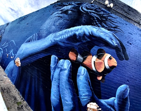 A vibrant mural of a clownfish being gently held by a large blue hand. The background features a monochromatic blue face with flowing hair. The artwork is painted on a brick wall and includes elements of the bricks incorporated as part of the design.